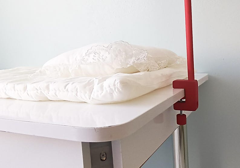 May include: A white quilted mattress pad with a decorative lace-trimmed pillow resting on top, secured to a white table surface by a red metal clamp mount with a vertical red pole extending upward from the side of the table edge.