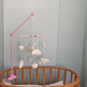 May include: A pink mobile arm attached to a wooden crib, featuring a hanging mobile with white plush shapes including stars, clouds, and a crescent moon suspended by white strings from a pink cross-shaped frame structure.