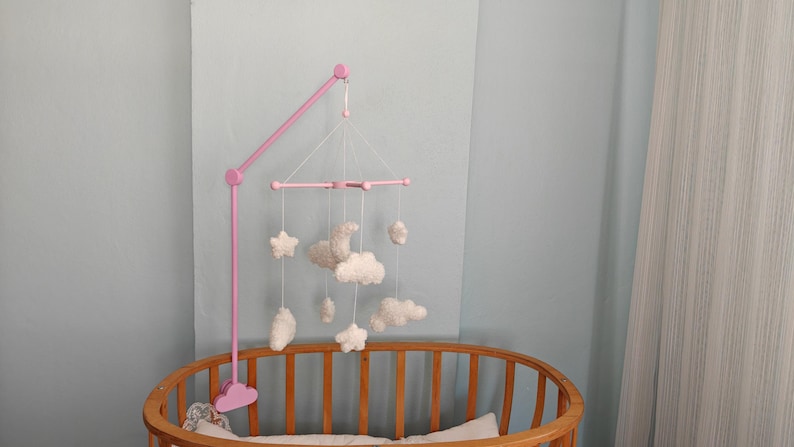 May include: A pink mobile arm attached to a wooden crib, featuring a hanging mobile with white plush shapes including stars, clouds, and a crescent moon suspended by white strings from a pink cross-shaped frame structure.