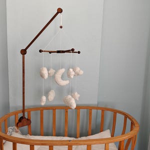 May include: A dark brown wooden mobile arm attached to a crib, featuring a cross-shaped hanger suspended by white strings. Hanging from the cross are several white, plush, cloud-shaped ornaments and a central white crescent moon ornament.