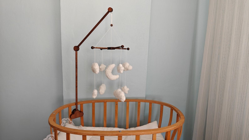 May include: A dark brown wooden mobile arm attached to a crib, featuring a cross-shaped hanger suspended by white strings. Hanging from the cross are several white, plush, cloud-shaped ornaments and a central white crescent moon ornament.