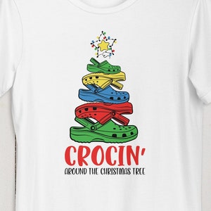 Könnte beinhalten: Weißes T-Shirt mit einem Weihnachtsbaum aus bunten Crocs. Der Baum ist mit einem Stern gekrönt und hat Lichter. Der Text "Crocin' around the Christmas tree" befindet sich unter dem Baum.