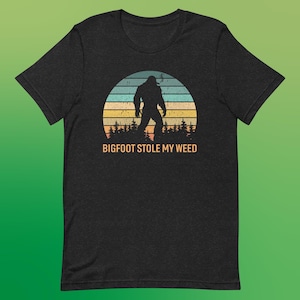 Op de afbeelding: Een zwart T-shirt met een retro-grafische afbeelding van Bigfoot die een joint rookt voor een zonsondergang. De tekst "BIGFOOT STOLE MY WEED" is onder de afbeelding gedrukt.
