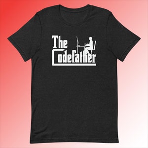 Op de afbeelding: Een zwart T-shirt met een wit grafisch ontwerp met de tekst "The Codefather" en een silhouet van een persoon die aan een computer zit.