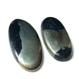 Pode incluir: Duas pedras de pirita ovais. As pedras têm um brilho metálico com áreas cinza escuro e dourado. As pedras são lisas e polidas, com uma aparência natural e orgânica. O fundo é branco.
