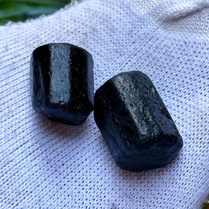 K&ouml;nnte beinhalten: Zwei schwarze, zylindrische Edelsteine mit rauer, strukturierter Oberfl&auml;che. Die Steine werden auf einem wei&szlig;en, strukturierten Stoff pr&auml;sentiert. Die Edelsteine werden wahrscheinlich f&uuml;r Schmuck oder Dekorationszwecke verwendet.