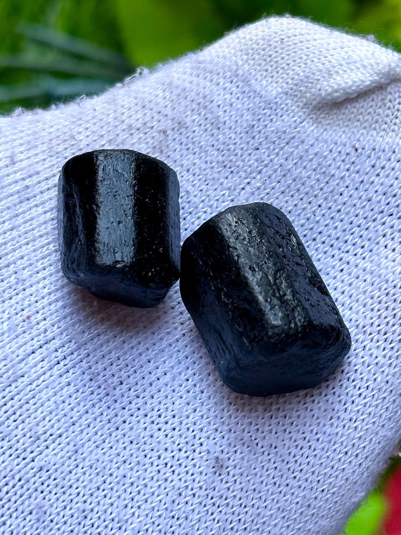 K&ouml;nnte beinhalten: Zwei schwarze, zylindrische Edelsteine mit rauer, strukturierter Oberfl&auml;che. Die Steine werden auf einem wei&szlig;en, strukturierten Stoff pr&auml;sentiert. Die Edelsteine werden wahrscheinlich f&uuml;r Schmuck oder Dekorationszwecke verwendet.