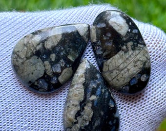 Puede incluir: Tres piedras pulidas en forma de lágrima con una mezcla de patrones grises, negros y azules. Las piedras se muestran sobre una superficie blanca texturizada, mostrando sus texturas y colores naturales. Probablemente gemas o elementos decorativos.