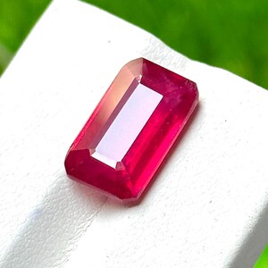 May include: A vibrant, rectangular-cut ruby gemstone with a deep red hue and faceted surfaces that reflect light. The stone features sharp, clean edges and a polished finish, showcasing a clear, transparent quality throughout its structure.