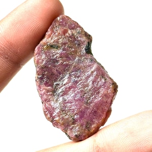 Puede incluir: Una gema de rubí en bruto con un tono rosa intenso, que muestra texturas y variaciones naturales. La piedra tiene una forma ligeramente irregular y una apariencia en bruto y sin pulir. El rubí se sostiene entre los dedos sobre un fondo blanco.