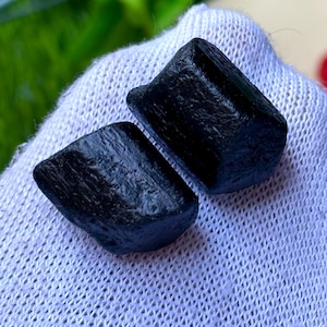 K&ouml;nnte beinhalten: Zwei grob geschnittene, schwarze Turmalinkristalle. Die Kristalle haben ein dunkles, undurchsichtiges Aussehen und eine leicht strukturierte Oberfl&auml;che. Sie werden auf einem wei&szlig;en, strukturierten Stoff pr&auml;sentiert, mit gr&uuml;nem Laub im Hintergrund. Die Kristalle sind tiefschwarz.