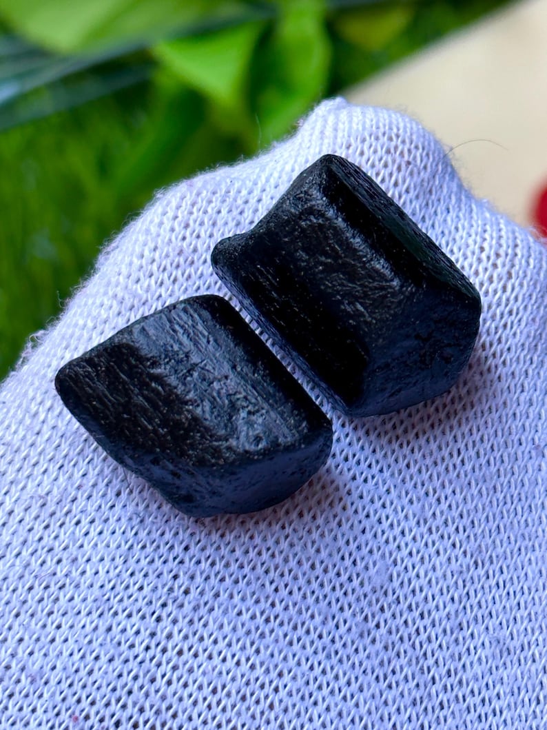 K&ouml;nnte beinhalten: Zwei grob geschnittene, schwarze Turmalinkristalle. Die Kristalle haben ein dunkles, undurchsichtiges Aussehen und eine leicht strukturierte Oberfl&auml;che. Sie werden auf einem wei&szlig;en, strukturierten Stoff pr&auml;sentiert, mit gr&uuml;nem Laub im Hintergrund. Die Kristalle sind tiefschwarz.