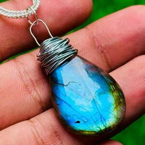 May include: A teardrop-shaped pendant with a vibrant blue and green iridescent gemstone. The stone is wrapped in silver wire and hangs from a silver chain. The pendant is held in a hand against a blurred green background.