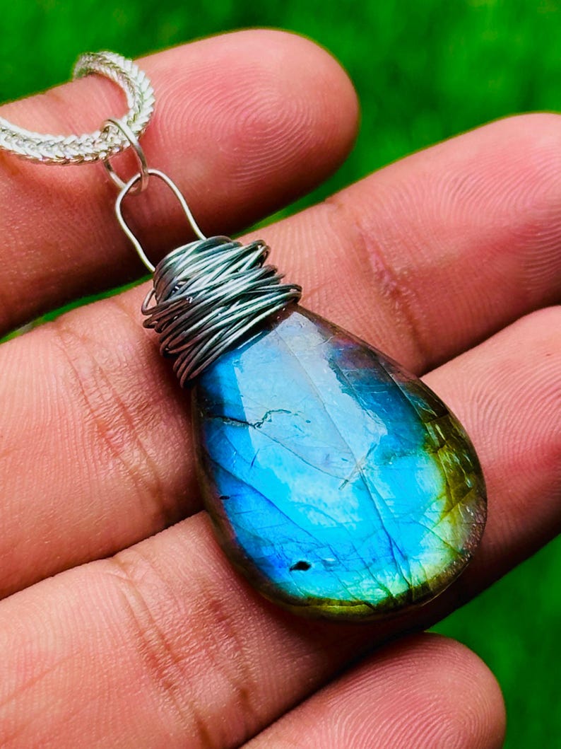May include: A teardrop-shaped pendant with a vibrant blue and green iridescent gemstone. The stone is wrapped in silver wire and hangs from a silver chain. The pendant is held in a hand against a blurred green background.