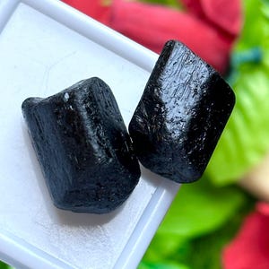 K&ouml;nnte beinhalten: Zwei rohe, schwarze Turmalinsteine liegen in einer wei&szlig;en Kunststoffschale. Die Steine haben eine raue, strukturierte Oberfl&auml;che und einen dunklen, gl&auml;nzenden Glanz. Der Hintergrund ist verschwommen, mit Andeutungen von Rot und Gr&uuml;n.