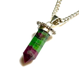 Könnte beinhalten: Eine silberne Halskette mit einem Anhänger aus einem grünen und weinroten Kristall. Der Kristall hat eine rechteckige Form mit spitzen Enden. Die Kette ist eine silberne Panzerkette.