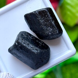 K&ouml;nnte beinhalten: Zwei rohe, schwarze Turmalinsteine liegen auf einem wei&szlig;en Tablett. Die Steine haben ein dunkles, fast obsidianartiges Aussehen mit sichtbarer Textur und nat&uuml;rlichen Unvollkommenheiten. Der Hintergrund ist verschwommen, mit Andeutungen von Rot und Gr&uuml;n.