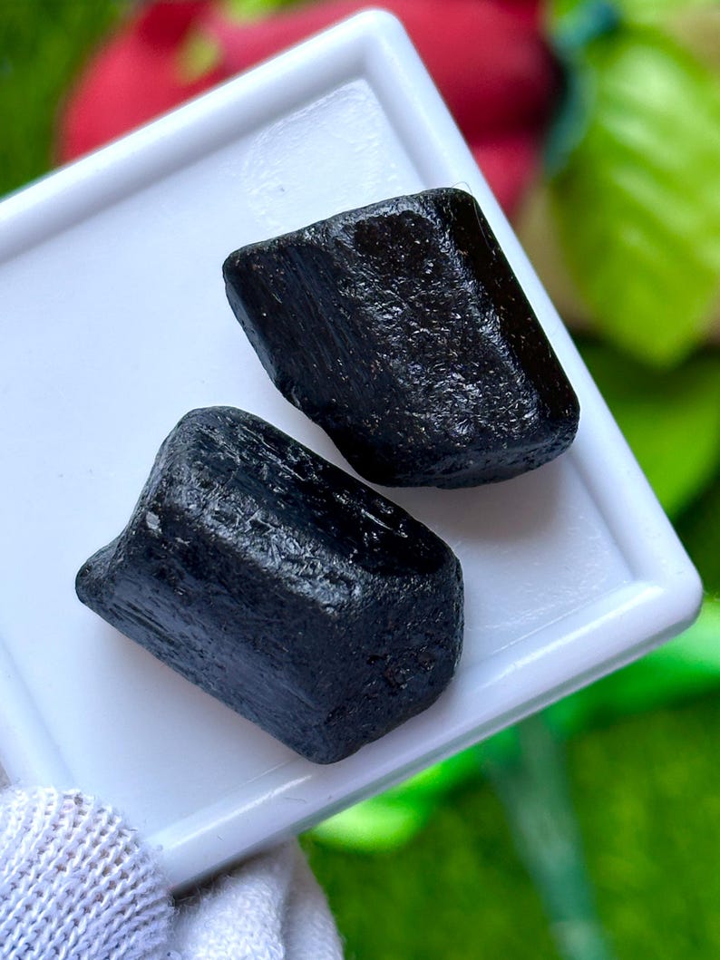 K&ouml;nnte beinhalten: Zwei rohe, schwarze Turmalinsteine liegen auf einem wei&szlig;en Tablett. Die Steine haben ein dunkles, fast obsidianartiges Aussehen mit sichtbarer Textur und nat&uuml;rlichen Unvollkommenheiten. Der Hintergrund ist verschwommen, mit Andeutungen von Rot und Gr&uuml;n.