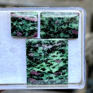 May include: Three square-cut zoisite gemstones with a mix of green, black, and pink hues. The stones are displayed in a clear plastic container, showcasing their natural patterns and colors. The gemstones vary in size, with the largest at the bottom.
