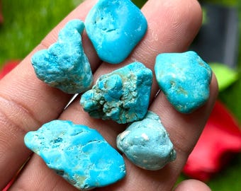 May include: A collection of turquoise gemstones in various shapes and sizes. The stones range in color from light to medium blue, with some showing natural matrix patterns. The gemstones are displayed against a blurred green and red background.