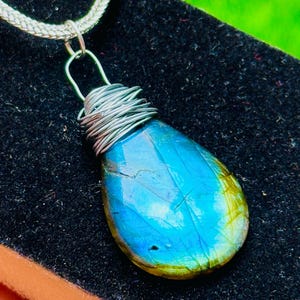 May include: A teardrop-shaped pendant with a vibrant blue and green gemstone, wrapped in silver wire. The pendant hangs from a silver chain, creating a striking contrast against the dark background. This unique piece is a beautiful accessory.
