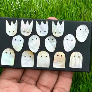 May include: A collection of 14 iridescent mother-of-pearl guitar fretboard inlays, arranged on a black surface. The inlays feature various shapes, including tulip and rounded designs, each with two small dot inlays.