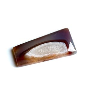Puede incluir: Una piedra de ágata rectangular con una superficie pulida. La piedra presenta bandas de tonos marrón, blanco y marrón rojizo. El centro tiene un patrón blanco cristalino y los bordes son de color marrón oscuro.