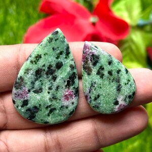 Może przedstawiać: Dwa kamienie szlachetne w kształcie łezki z zieloną podstawą, nakrapiane czernią i różowymi plamkami. Kamienie trzymane są w dłoni, z rozmytym tłem zieleni i czerwoną kwiatem.