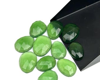 Op de afbeelding: Een verzameling gefacetteerde, ovaalvormige groene edelstenen. De edelstenen variëren in groentinten, van licht tot donker, en worden gepresenteerd tegen een witte achtergrond met een zwarte bak in de hoek. De edelstenen zijn waarschijnlijk voor het maken van sieraden.