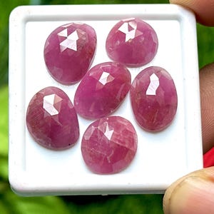 May include: Five oval-shaped, faceted pink rubies arranged on a white square display tray. Each gemstone features a rose-cut surface with triangular facets that reflect light, showcasing a deep magenta hue and a translucent, polished appearance.