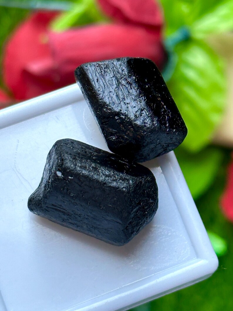 K&ouml;nnte beinhalten: Zwei rohe, schwarze Turmalinkristalle mit rauer, strukturierter Oberfl&auml;che. Die Kristalle werden auf einem wei&szlig;en Tablett mit einem verschwommenen roten und gr&uuml;nen Hintergrund pr&auml;sentiert.