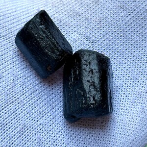 K&ouml;nnte beinhalten: Zwei rohe, schwarze Turmalinkristalle. Die Kristalle haben eine raue, strukturierte Oberfl&auml;che und eine dunkle, undurchsichtige Farbe. Sie werden auf einem wei&szlig;en, strukturierten Stoff pr&auml;sentiert.