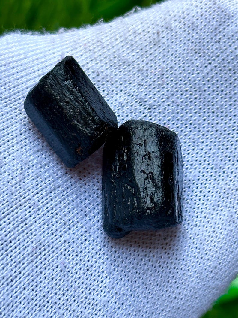 K&ouml;nnte beinhalten: Zwei rohe, schwarze Turmalinkristalle. Die Kristalle haben eine raue, strukturierte Oberfl&auml;che und eine dunkle, undurchsichtige Farbe. Sie werden auf einem wei&szlig;en, strukturierten Stoff pr&auml;sentiert.