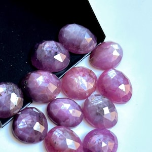 Peut inclure: Une collection de pierres précieuses ovales et facettées, dans des tons de violet et de rose. Les gemmes ont une surface polie et réfléchissante, avec de multiples facettes captant la lumière. Les pierres sont disposées sur une surface blanche avec un bord noir et blanc incliné.