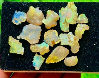 Puede incluir: Una colección de piedras preciosas de ópalo en bruto, que muestra una variedad de formas y tamaños. Los ópalos exhiben una mezcla de colores, incluyendo blanco lechoso, amarillo y destellos de azul y verde. Las piedras se muestran sobre una superficie negra.
