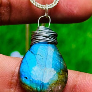 May include: A teardrop-shaped pendant with a vibrant blue and green gemstone. The stone is wrapped in silver wire and suspended from a silver chain. The gemstone has a polished surface and displays a range of colors, including blues, greens, and hints of gold.