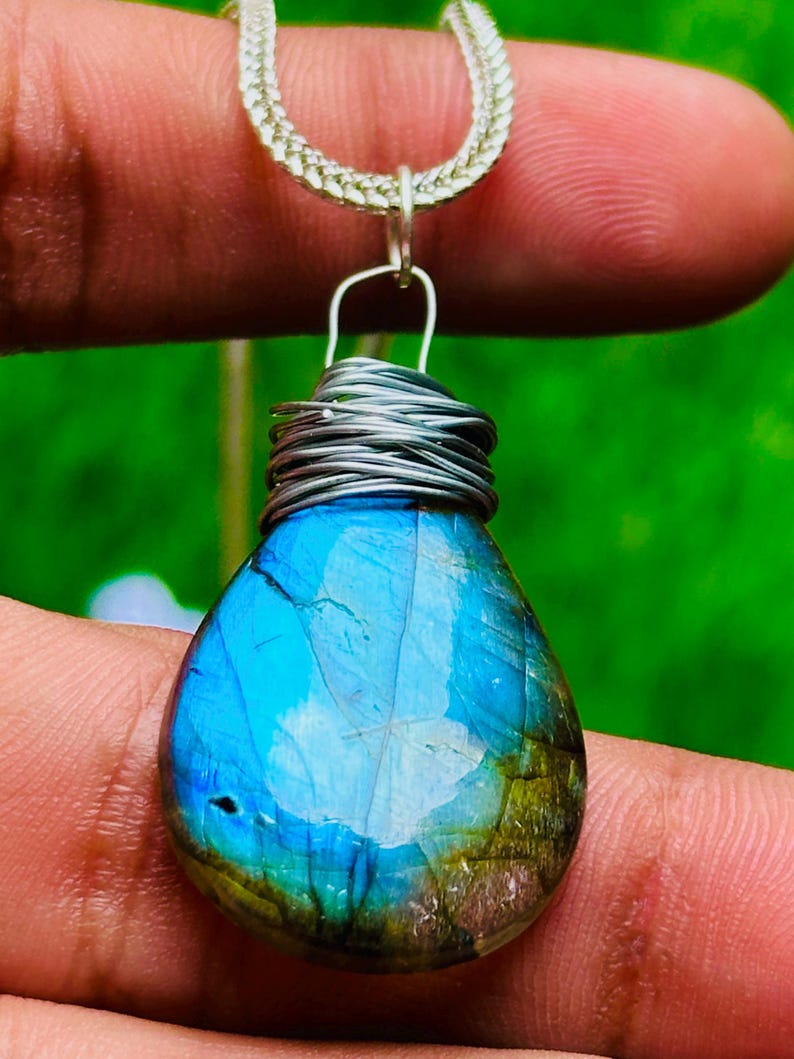 May include: A teardrop-shaped pendant with a vibrant blue and green gemstone. The stone is wrapped in silver wire and suspended from a silver chain. The gemstone has a polished surface and displays a range of colors, including blues, greens, and hints of gold.