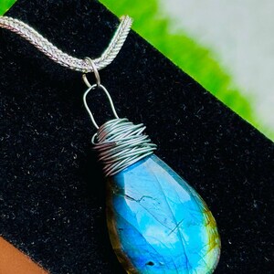 May include: A teardrop-shaped labradorite pendant with a silver wire wrap and chain. The gemstone displays a vibrant blue and green iridescence. The pendant hangs from a delicate silver chain, creating a striking piece of jewelry.