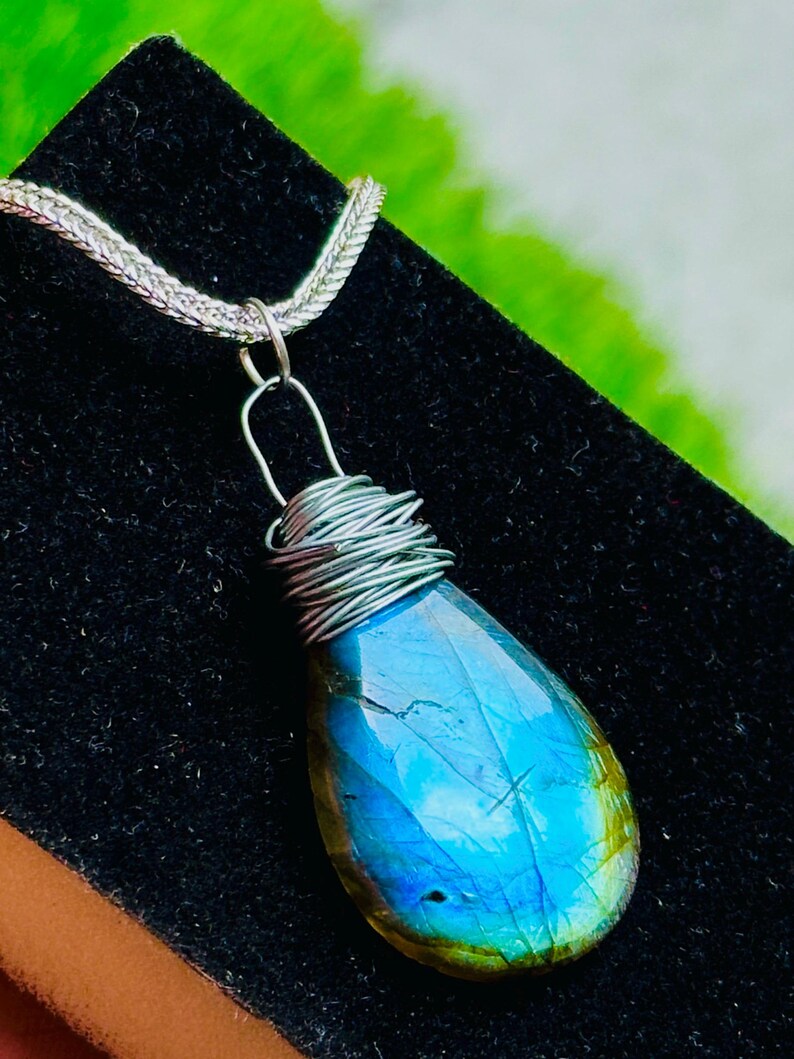 May include: A teardrop-shaped labradorite pendant with a silver wire wrap and chain. The gemstone displays a vibrant blue and green iridescence. The pendant hangs from a delicate silver chain, creating a striking piece of jewelry.