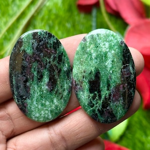 Può includere: Due pietre ovali di zoisite, con una miscela di verde, nero e bordeaux. Le pietre lucide risaltano su uno sfondo sfocato verde e rosso, ideali per la creazione di gioielli.