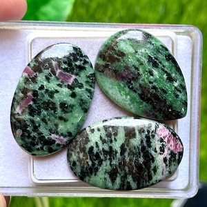 Puede incluir: Tres piedras preciosas de zoisita pulida con inclusiones de rubí. Las piedras, de forma ovalada y de lágrima, muestran un patrón moteado de tonos verdes, negros y rosas. Se presentan en un recipiente de plástico transparente, con un fondo verde borroso.
