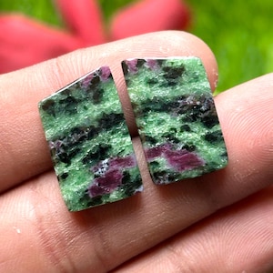 Puede incluir: Dos piedras preciosas rectangulares con un patrón moteado verde y negro y parches de color rojo intenso. Las piedras se sostienen en una mano contra un fondo verde y rojo borroso. Para la creación de joyas.