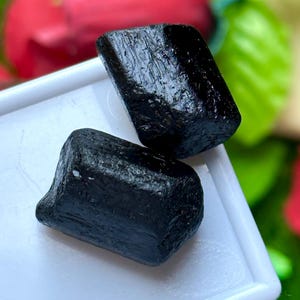 K&ouml;nnte beinhalten: Zwei rohe, schwarze Turmalinkristalle mit rauer, strukturierter Oberfl&auml;che. Die Kristalle werden auf einem wei&szlig;en Tablett mit einem verschwommenen roten und gr&uuml;nen Hintergrund pr&auml;sentiert.