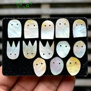 May include: A collection of 13 iridescent mother-of-pearl pieces in various shapes, including rounded rectangles, crown-like designs, and ovals. Each piece has two small drilled holes, resembling eyes. The pieces are arranged on a black surface.