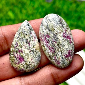 Può includere: Due pietre preziose lucide, una a goccia e una ovale, tenute in mano. Presentano un mix di bianco, verde, nero e magenta. Lo sfondo è verde sfocato. Adatto per la creazione di gioielli.