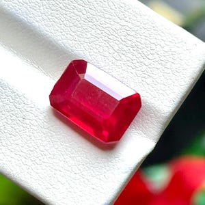 Puede incluir: Una gema de rubí facetada y rectangular, de un intenso color rojo. La gema se exhibe sobre una superficie blanca texturizada, mostrando su claridad y brillo. El corte de la piedra resalta sus facetas, reflejando la luz y creando un efecto brillante.