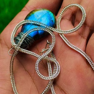 May include: A silver necklace with a blue and green labradorite pendant. The pendant is wrapped in silver wire and is suspended from a woven chain. The labradorite has a vibrant blue iridescence.