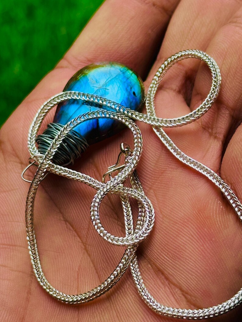 May include: A silver necklace with a blue and green labradorite pendant. The pendant is wrapped in silver wire and is suspended from a woven chain. The labradorite has a vibrant blue iridescence.