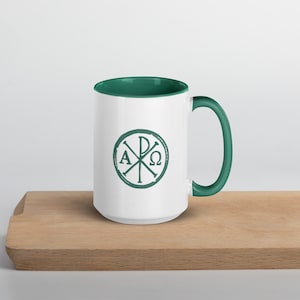 Peut inclure: Une tasse en céramique blanche avec un bord et une poignée verts. La tasse présente un motif vert du symbole Chi Rho, un symbole chrétien qui représente le Christ. Le symbole est entouré d'un cercle avec les lettres Alpha et Omega.