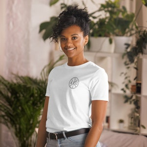 Può includere: Una maglietta bianca con un design circolare grigio che presenta le lettere greche Alfa e Omega, circondate da una croce.